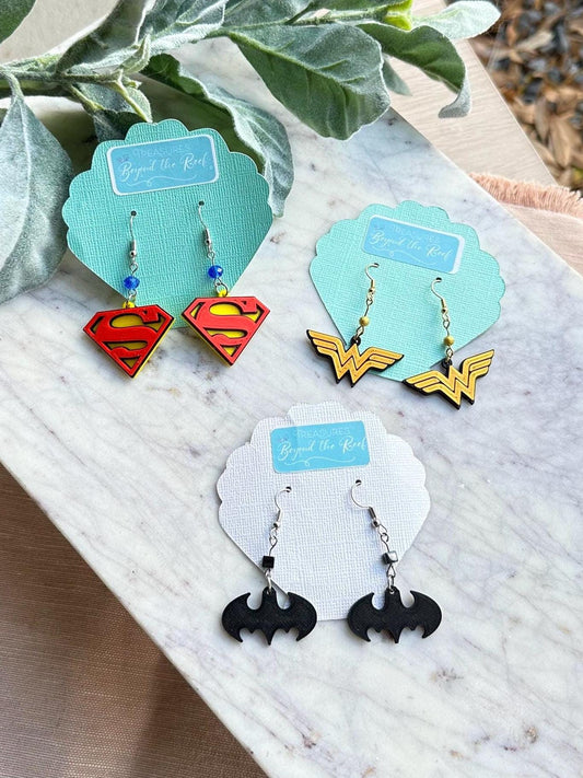 May include: Three pairs of superhero themed earrings on white and blue display cards. The earrings are silver with black, red, yellow, and blue accents. The cards have the words 'Treasures Beyond the Reef' printed on them.