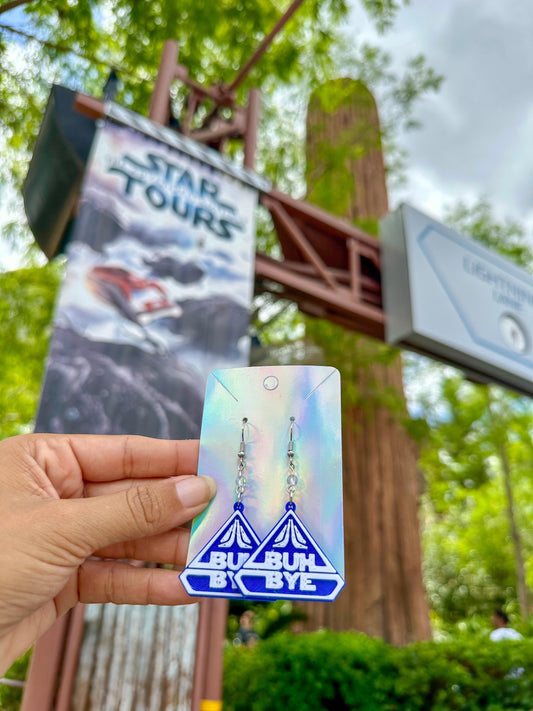 May include: A pair of blue and white earrings with the words 'BUH BYE' on them. The earrings are hanging from a silver hook.