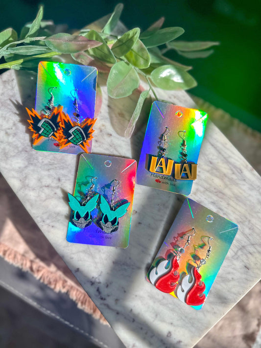 May include: Four pairs of earrings on display cards. The earrings are made of colorful resin and feature different characters and designs. The earrings are handmade with love. The earrings are displayed on iridescent cards with the words 'Handmade with love' printed on them.