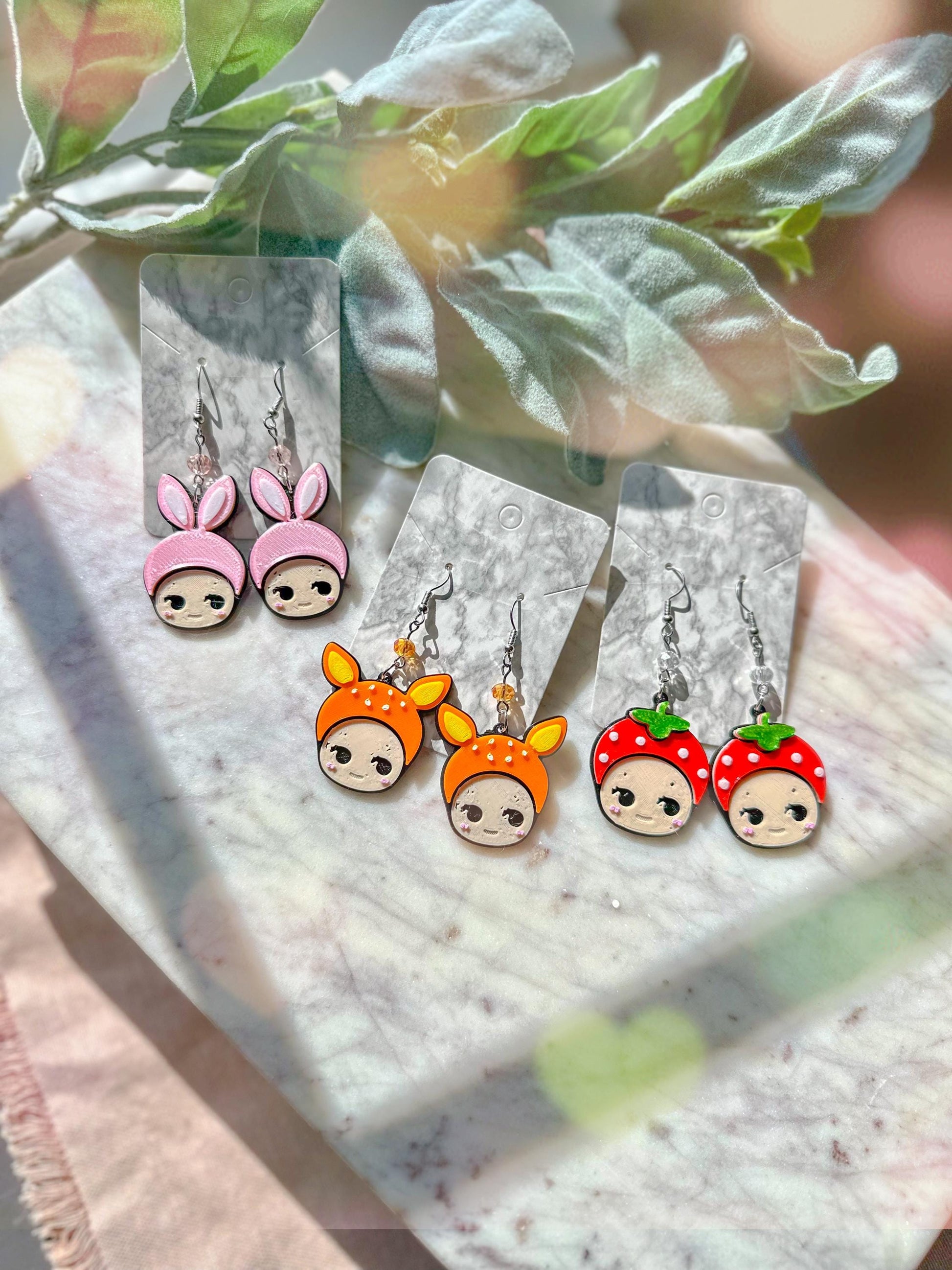 May include: Three pairs of dangle earrings with cartoon faces. The first pair has pink bunny ears, the second pair has orange fox ears, and the third pair has red strawberry tops. Each earring has a silver hook and a small silver bead.
