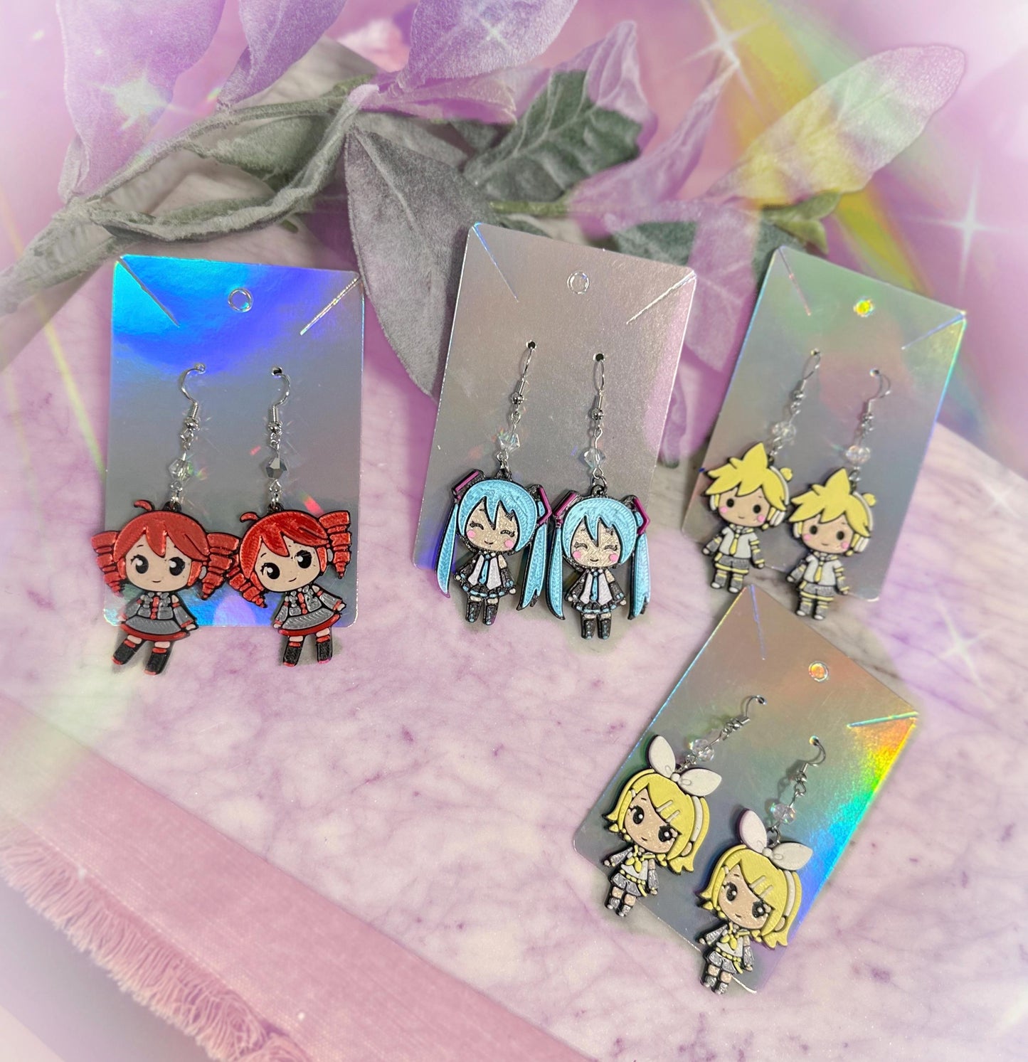 May include: Earrings featuring anime-style characters. The earrings are on silver cards. One pair features red-haired characters, another pair has blue-haired characters, a third pair has yellow-haired characters, and the last pair has yellow-haired characters with white bows. The earrings are made of a shiny material.