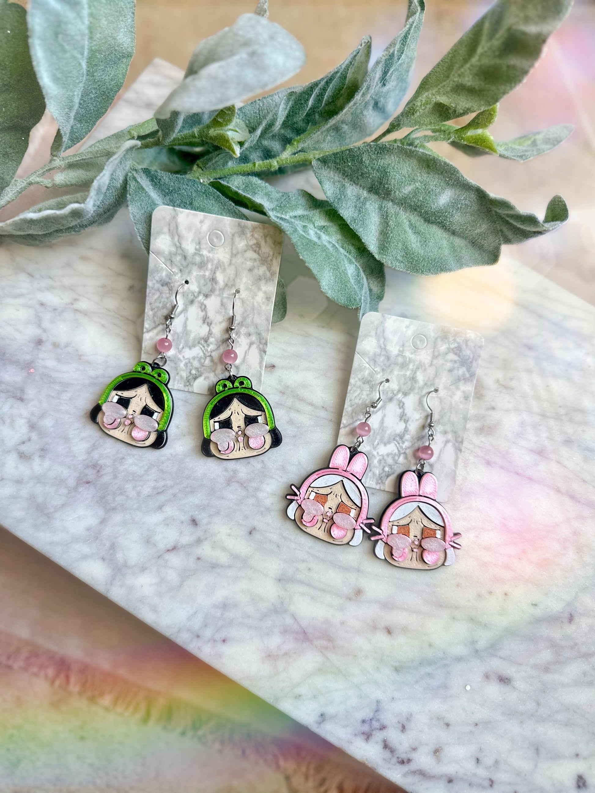 May include: Two pairs of dangle earrings. One pair features a green cartoon character with pink cheeks and a pink bow. The other pair features a pink cartoon character with pink cheeks and a pink bow.